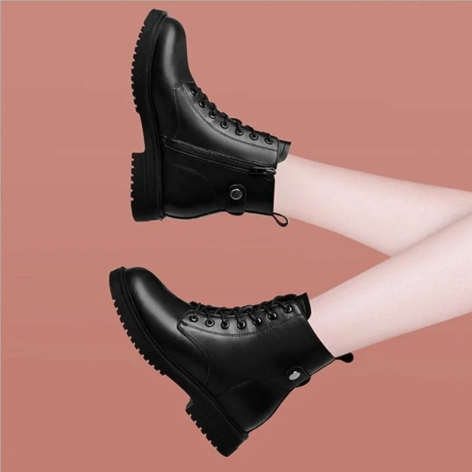 Side profile of black ankle boots suspended against a pink background, highlighting the lace-up fronts, side zipper panels, pull tabs, and lugged soles.
