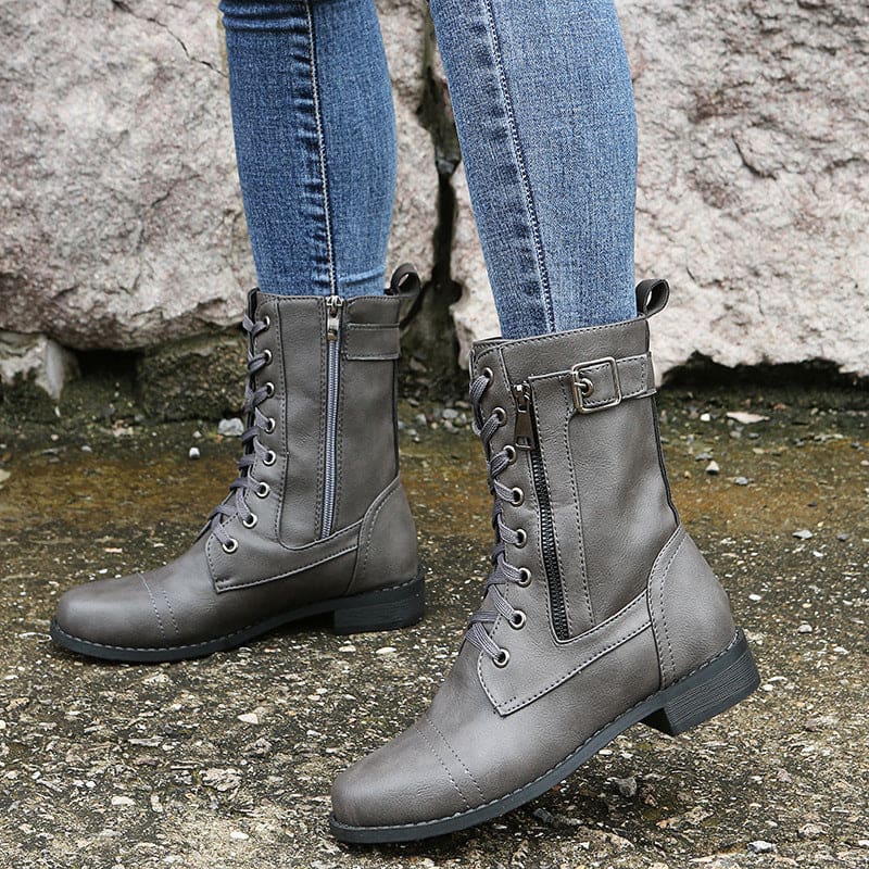 Pair of lace-up combat boots with side zipper and buckle detail worn with jeans on a stone background._10