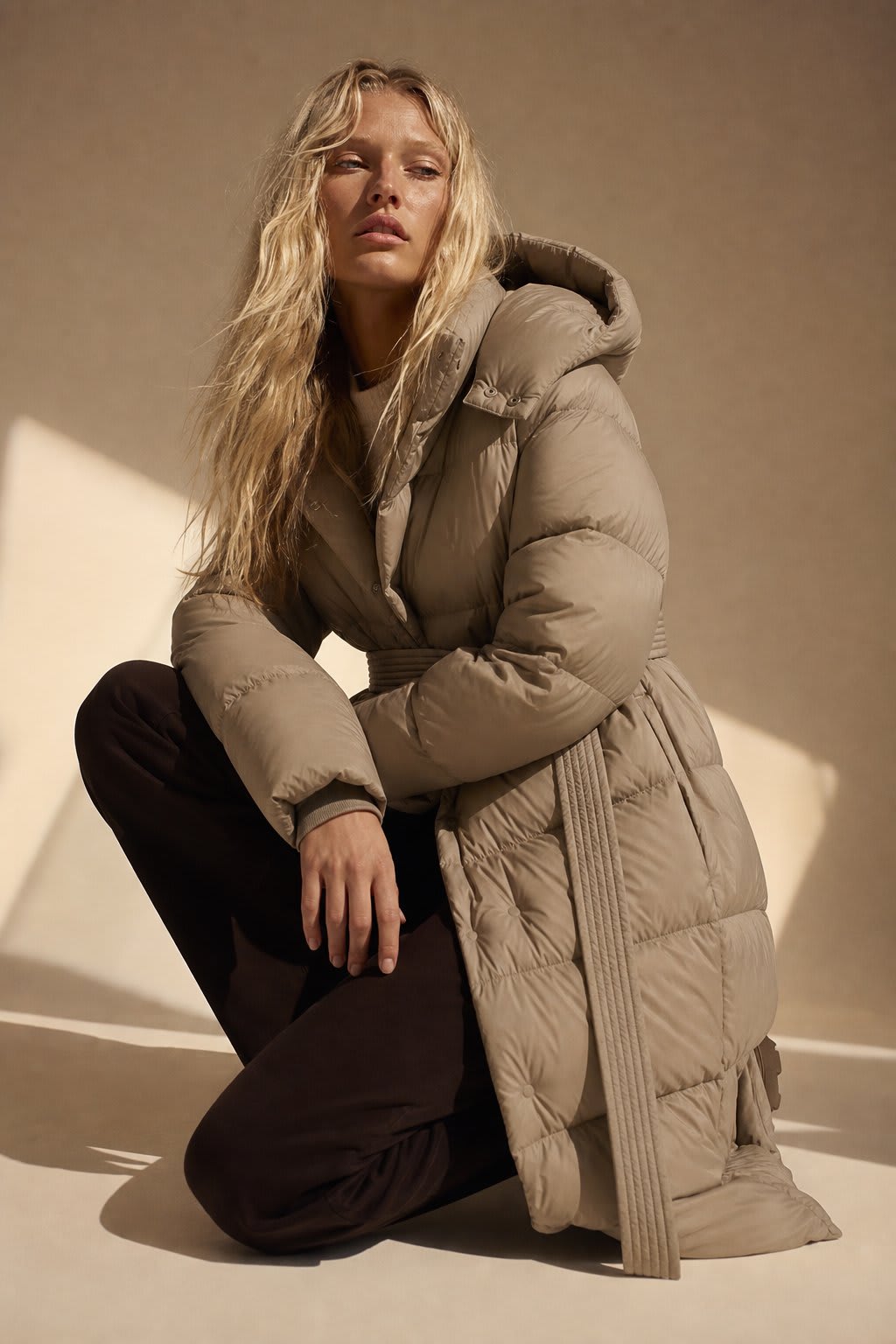 Woman wearing a long belted puffer coat with an oversized hood in a neutral tone, crouching against a minimalist beige background._0