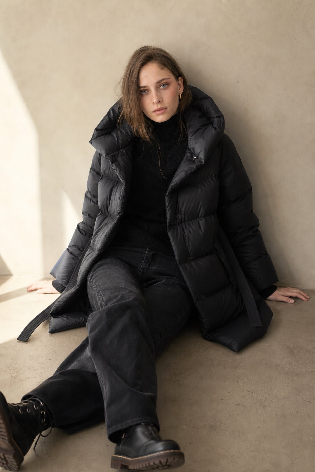 Woman wearing a long belted puffer coat with an oversized hood in a neutral tone, crouching against a minimalist beige background._2