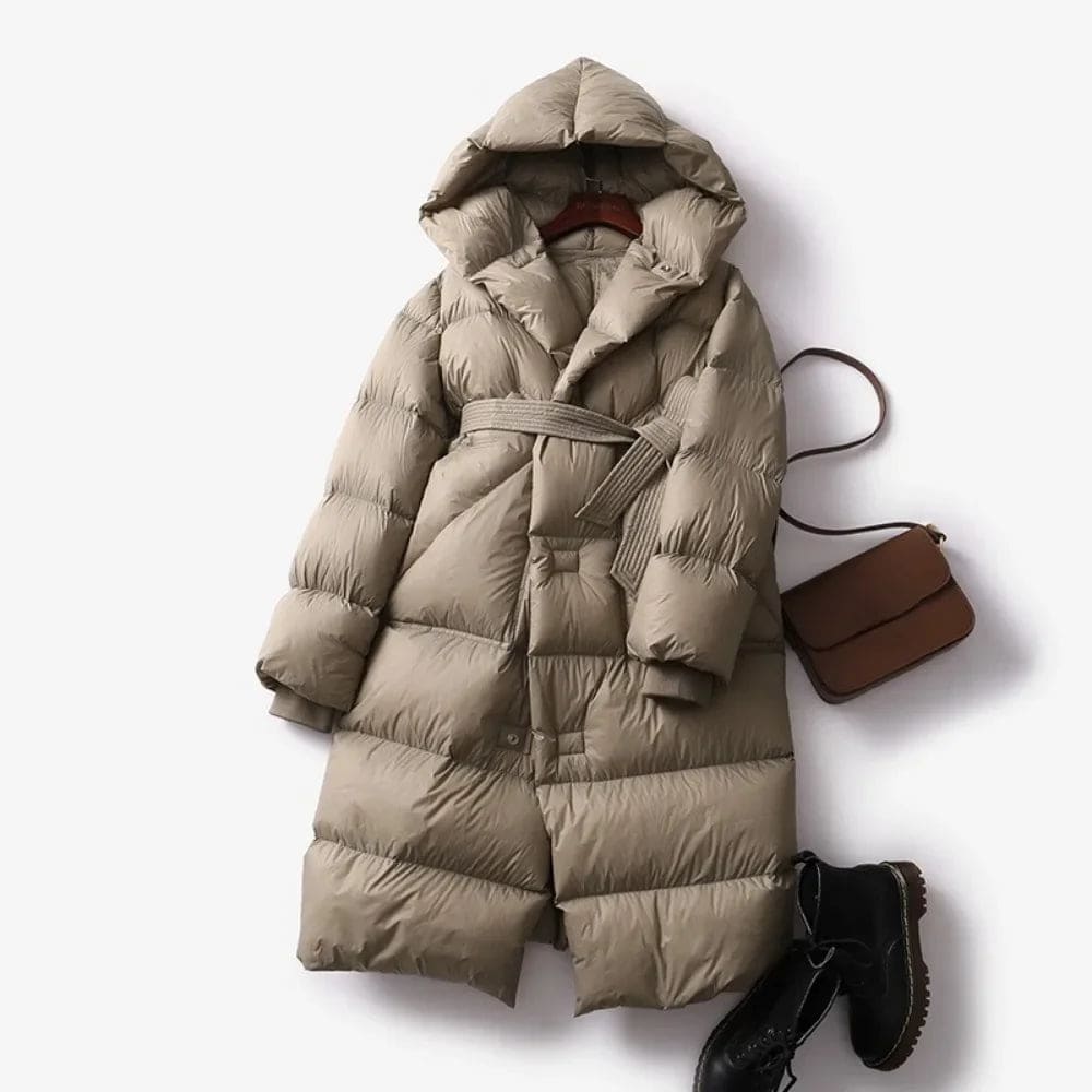 Woman wearing a long belted puffer coat with an oversized hood in a neutral tone, crouching against a minimalist beige background._4