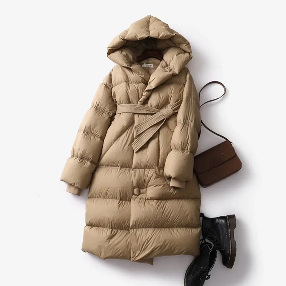 Woman wearing a long belted puffer coat with an oversized hood in a neutral tone, crouching against a minimalist beige background._5