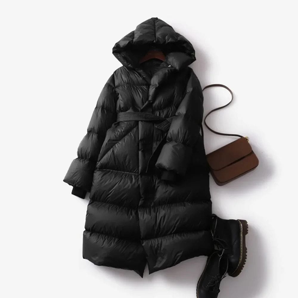 Woman wearing a long belted puffer coat with an oversized hood in a neutral tone, crouching against a minimalist beige background._6