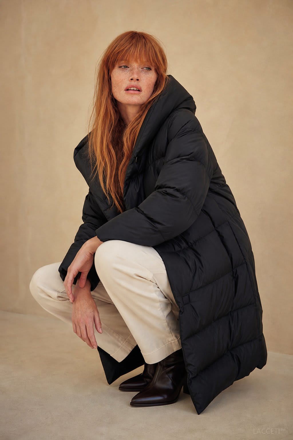 Model wearing a long black puffer parka with oversized fit and structured hood, styled with cream trousers and black ankle boots_0