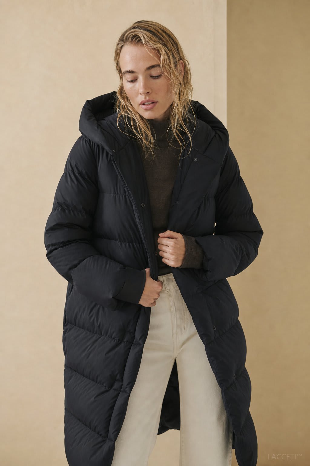 Model wearing a long black puffer parka with oversized fit and structured hood, styled with cream trousers and black ankle boots_4