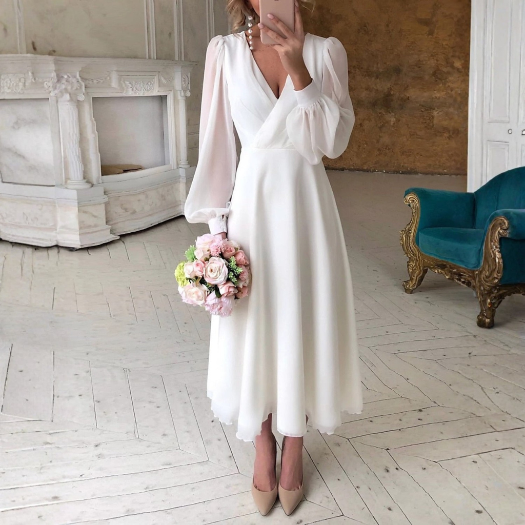 Woman wearing a white long sleeve midi wedding dress with V-neck and flowing skirt, holding a bouquet of pink flowers in an elegant room._0