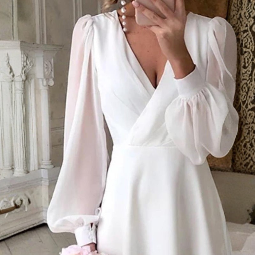 Woman wearing a white long sleeve midi wedding dress with V-neck and flowing skirt, holding a bouquet of pink flowers in an elegant room._1