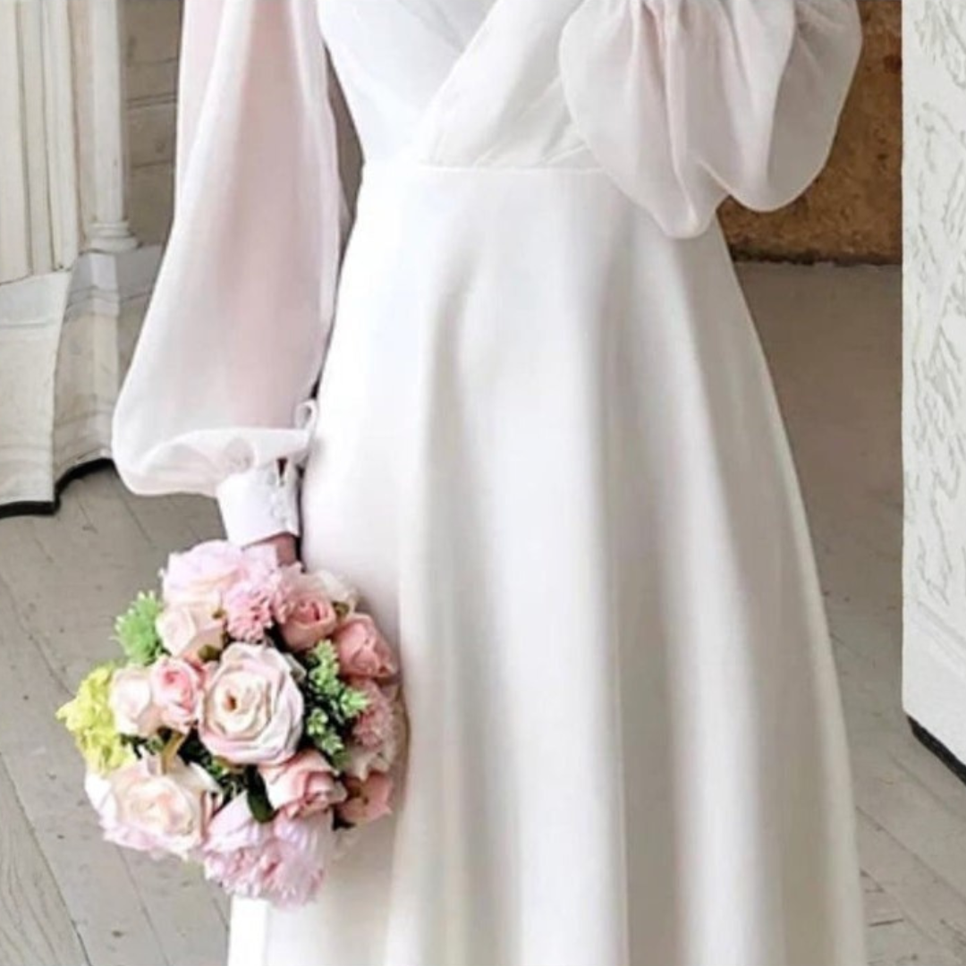 Woman wearing a white long sleeve midi wedding dress with V-neck and flowing skirt, holding a bouquet of pink flowers in an elegant room._2