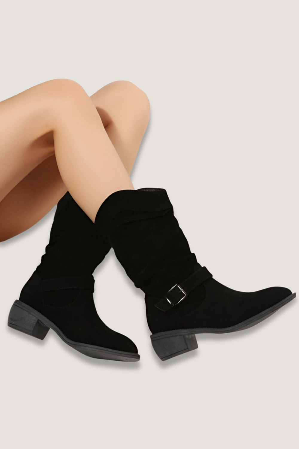 Black mid-calf boots crossed over a seated model’s legs, showing pointed toes, buckle straps, and low block heels.