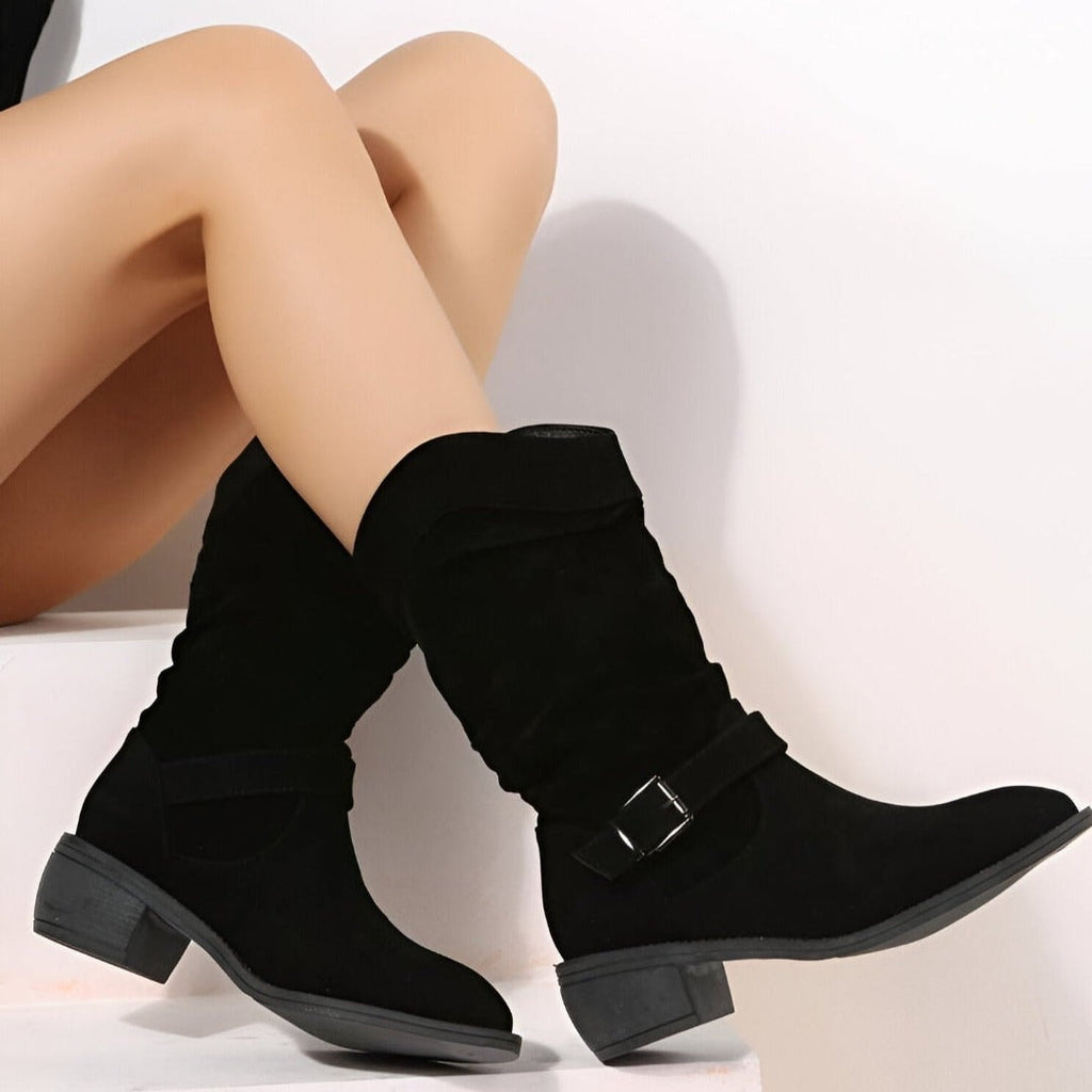 Black boots worn with crossed legs against a white background, highlighting the buckle strap and angled side profile.