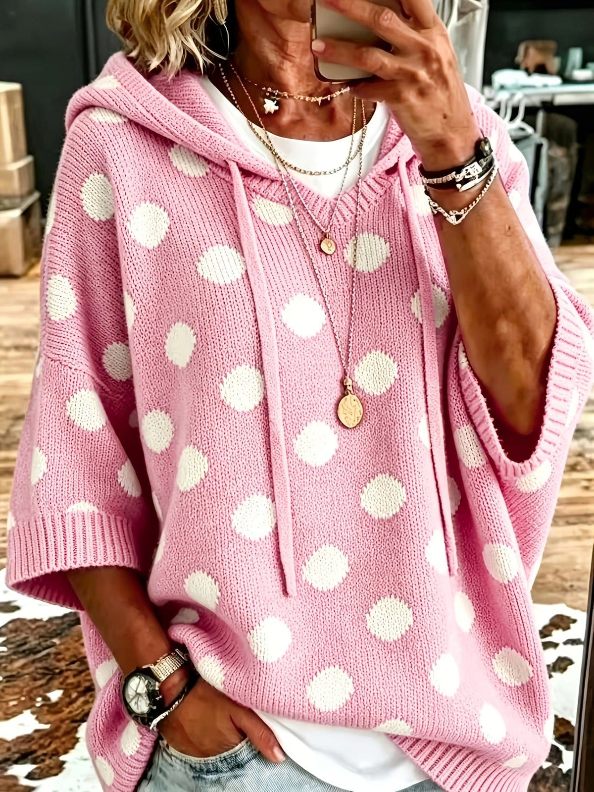 Person wearing a loose pink hooded knit pullover with white polka dots, drawstring ties and half sleeves over a white top and jeans._0