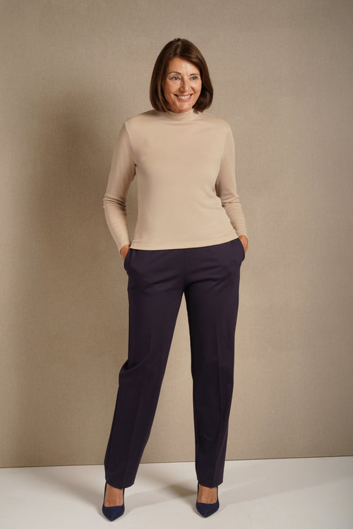 Woman wearing a beige long sleeve mock neck top with dark purple straight leg trousers and navy heels._0
