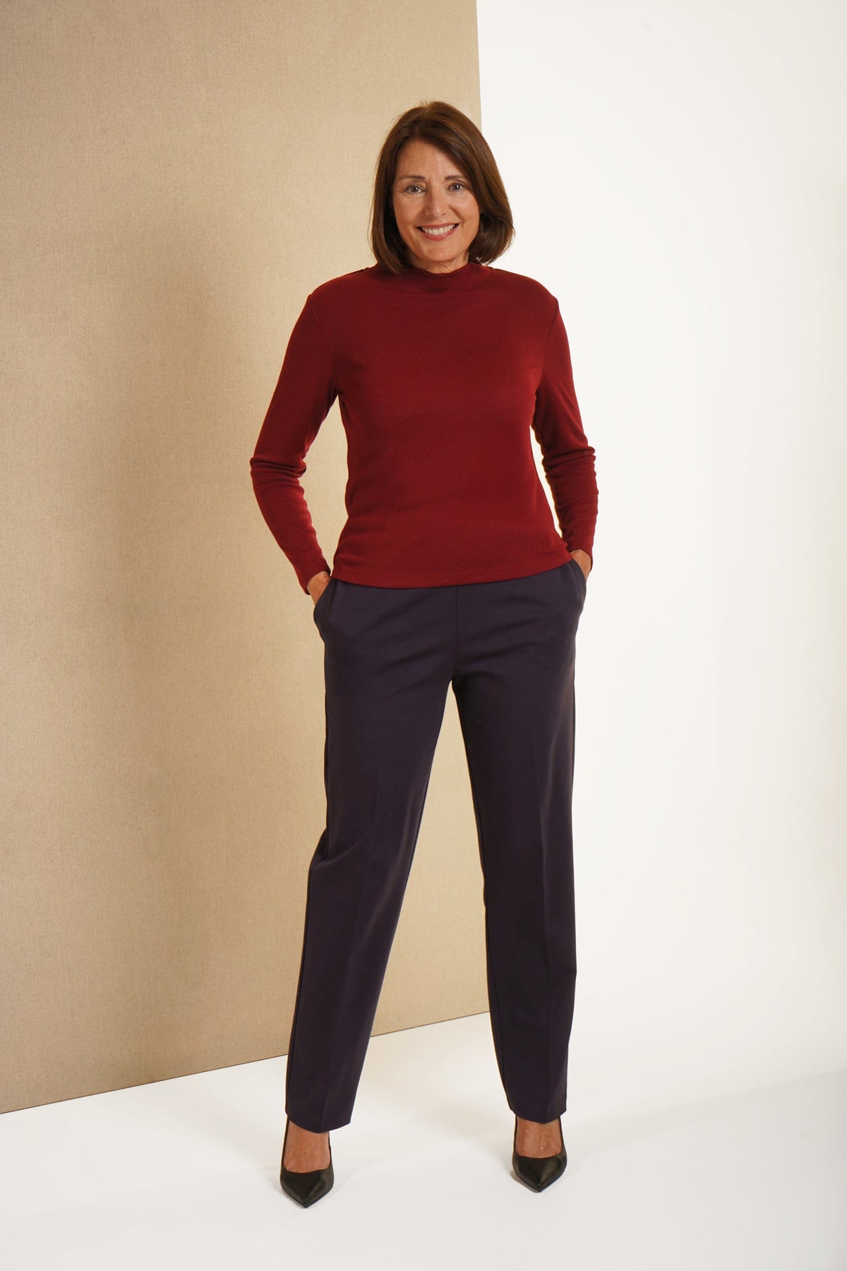 Woman wearing a beige long sleeve mock neck top with dark purple straight leg trousers and navy heels._2
