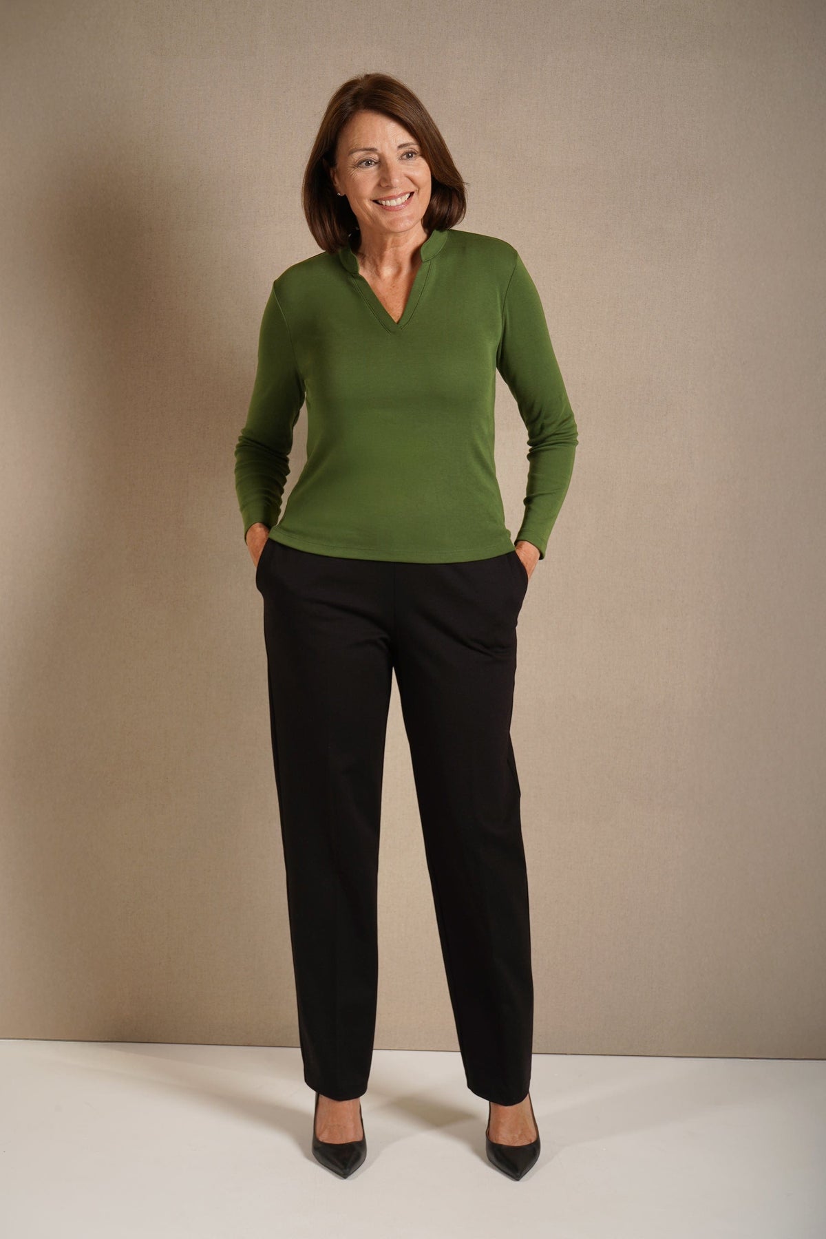Woman wearing a beige long sleeve mock neck top with dark purple straight leg trousers and navy heels._3