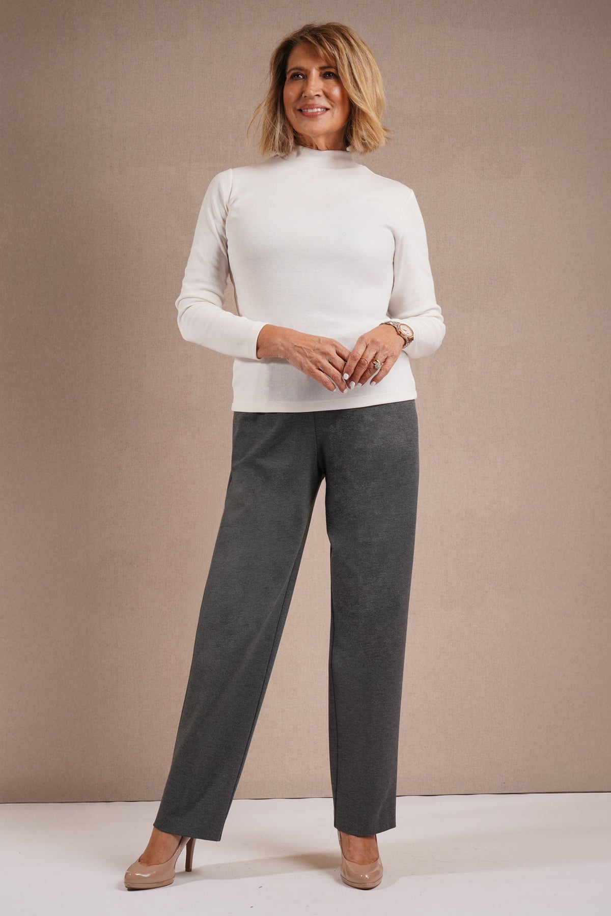 Woman wearing a beige long sleeve mock neck top with dark purple straight leg trousers and navy heels._4