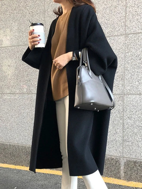 Black open-front longline coat worn over a camel top and light trousers, with a structured gray handbag at the side.