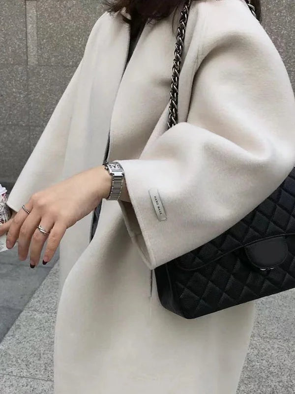 Close view of the light cream coat highlighting its open front, rounded bracelet sleeve, and sleeve label beside a quilted black handbag.