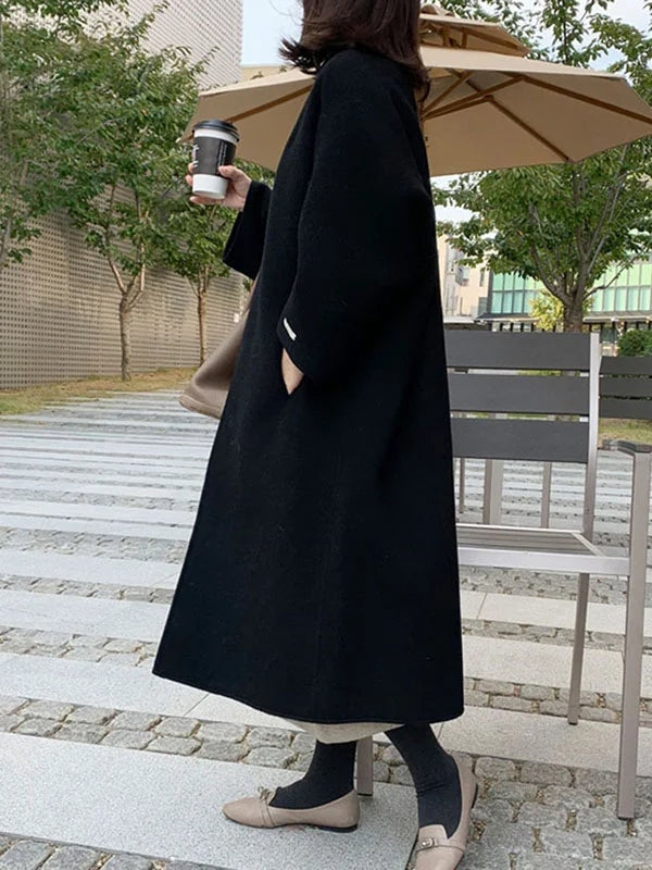 Back and side view of the black coat revealing its longline length, loose silhouette, and bracelet-length sleeve in an outdoor setting.