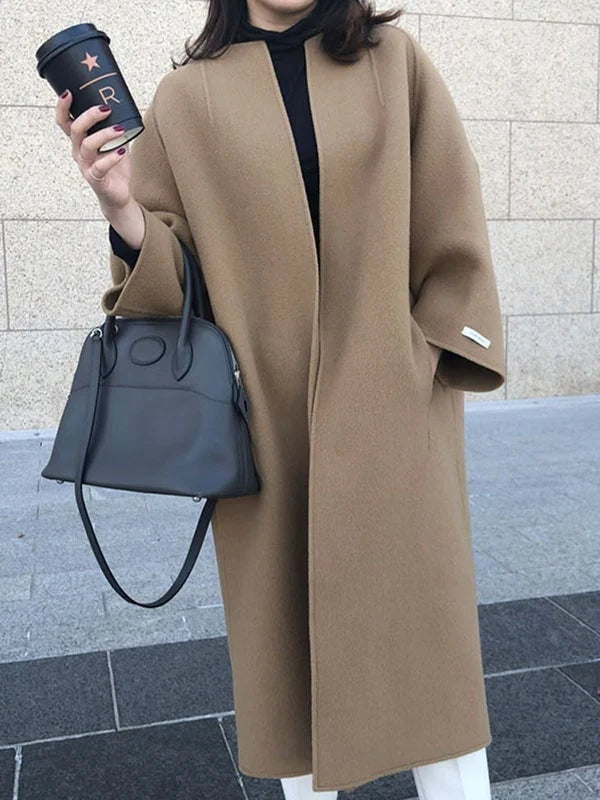 Front view of the camel coat showing its open front, longline cut, bracelet sleeves, and curved hem over white trousers.