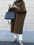 Three-quarter view of the dark brown longline coat with folded bracelet sleeves, worn over white trousers and paired with a black handbag.