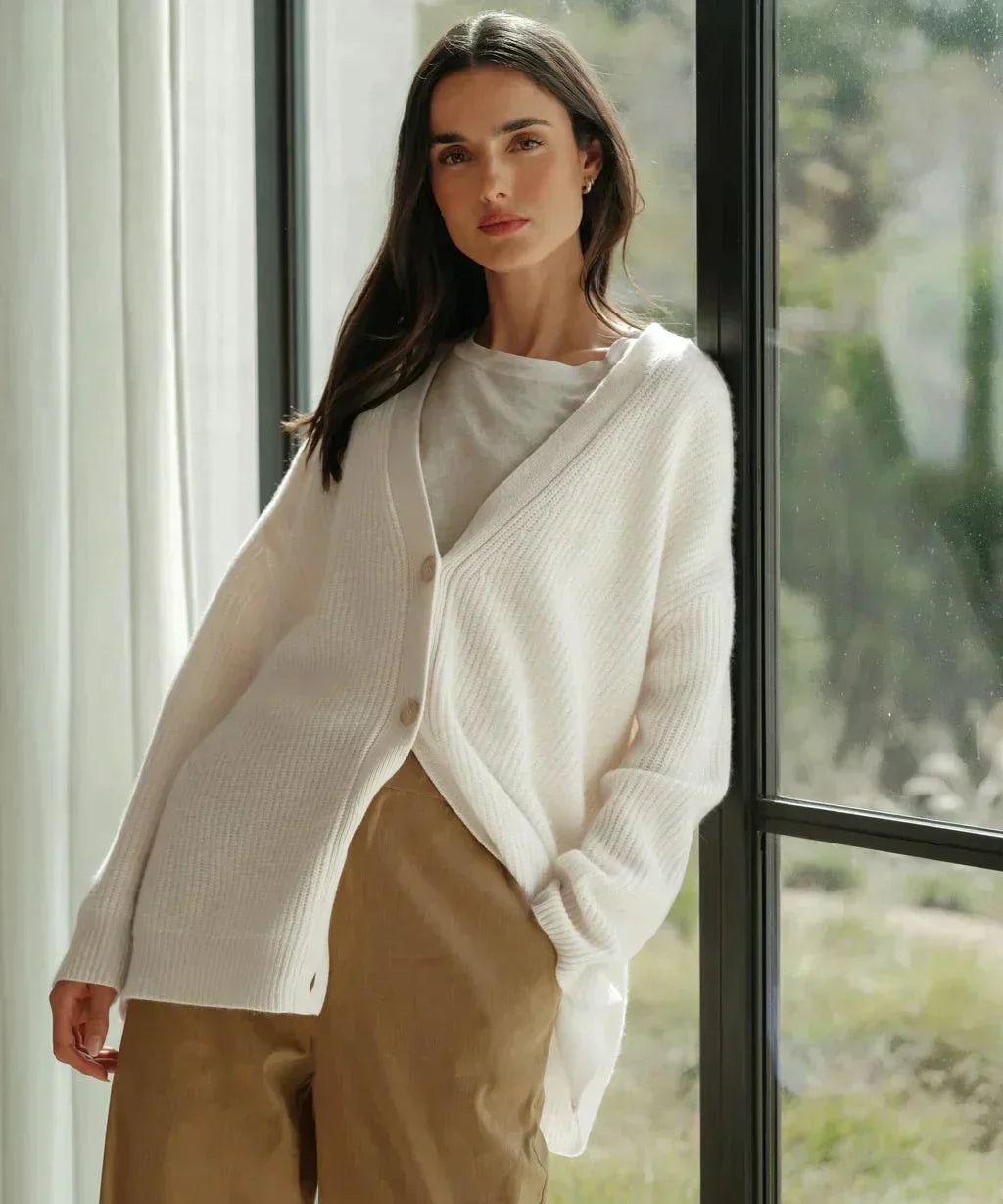 Cream oversized V-neck cardigan worn partly buttoned over a white top and tan trousers beside a window.