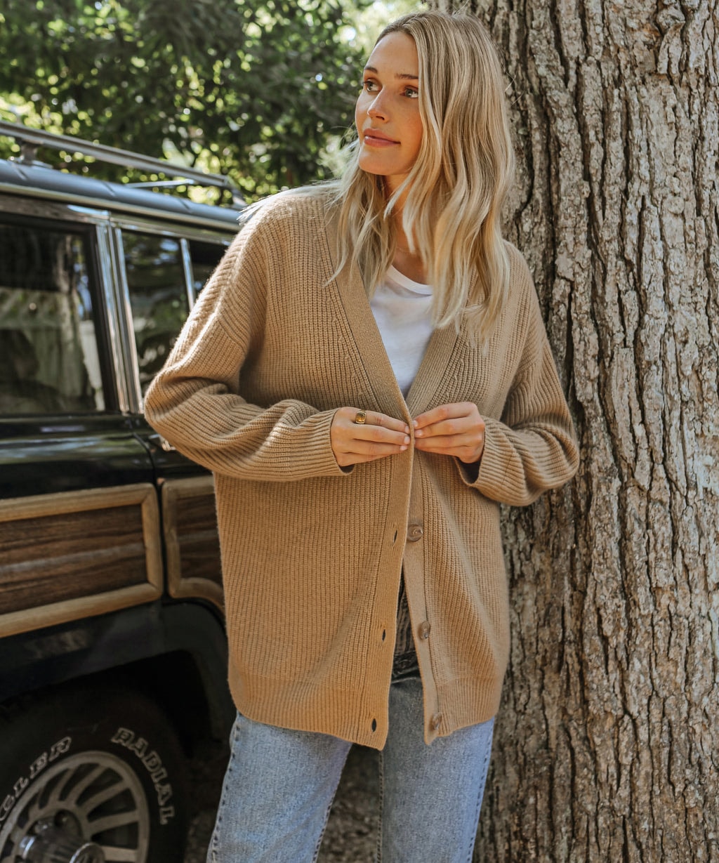 Camel oversized V-neck cardigan being buttoned over a white T-shirt and blue jeans outdoors.