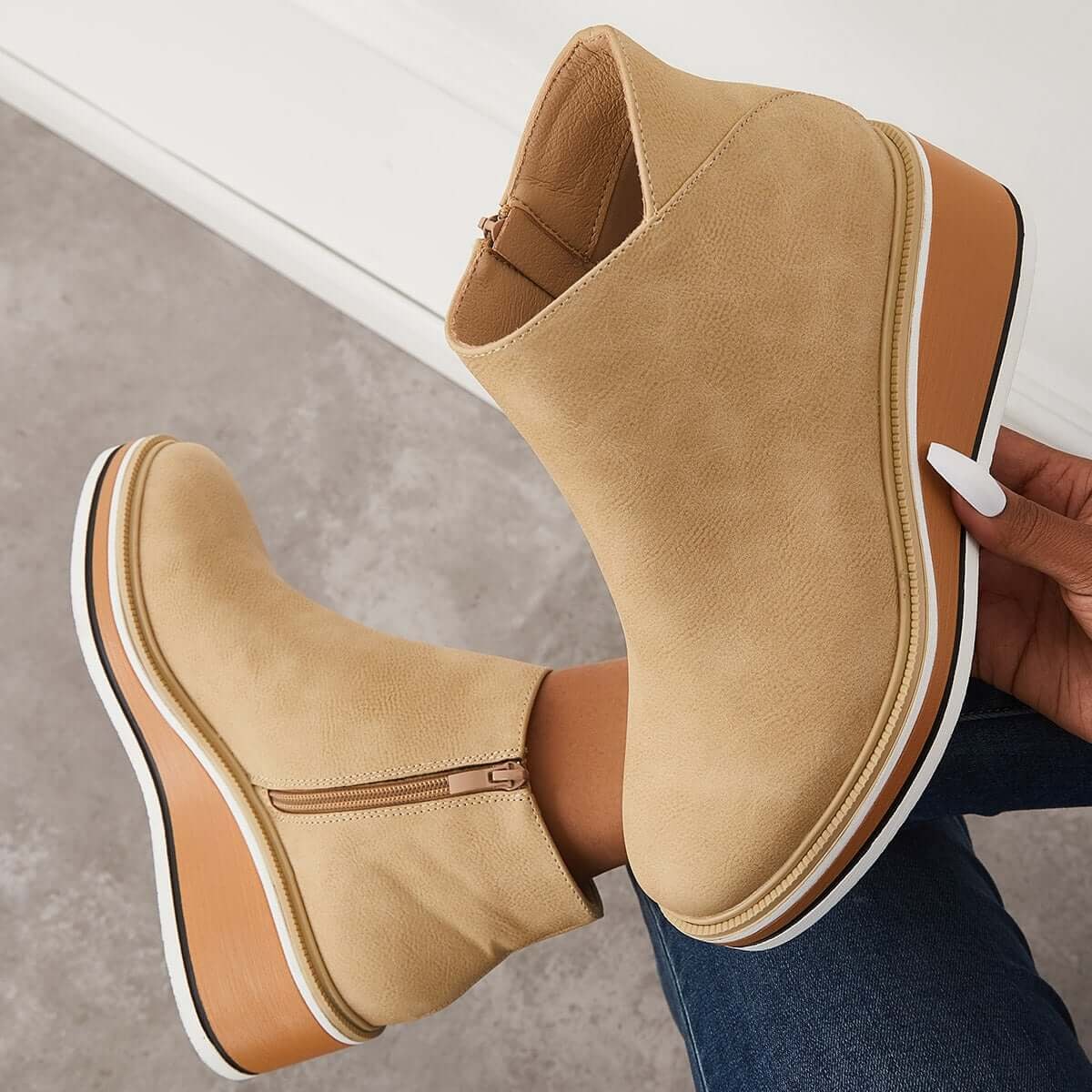 Light beige ankle boot held above another boot, displaying the side zipper and layered wedge sole.