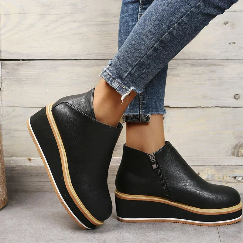 Black platform ankle boots shown at an angle against a light wood backdrop, with one boot lifted.