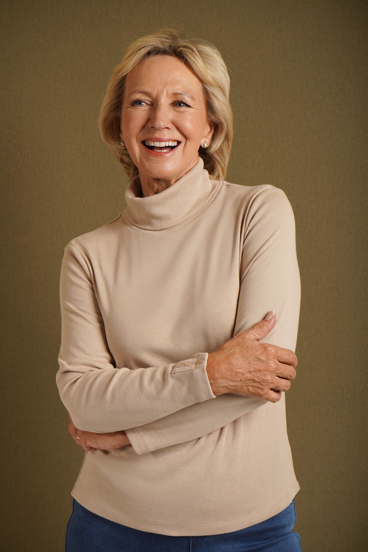 Woman wearing a beige long sleeve turtleneck top with buttoned cuffs and blue jeans against an olive backdrop._0