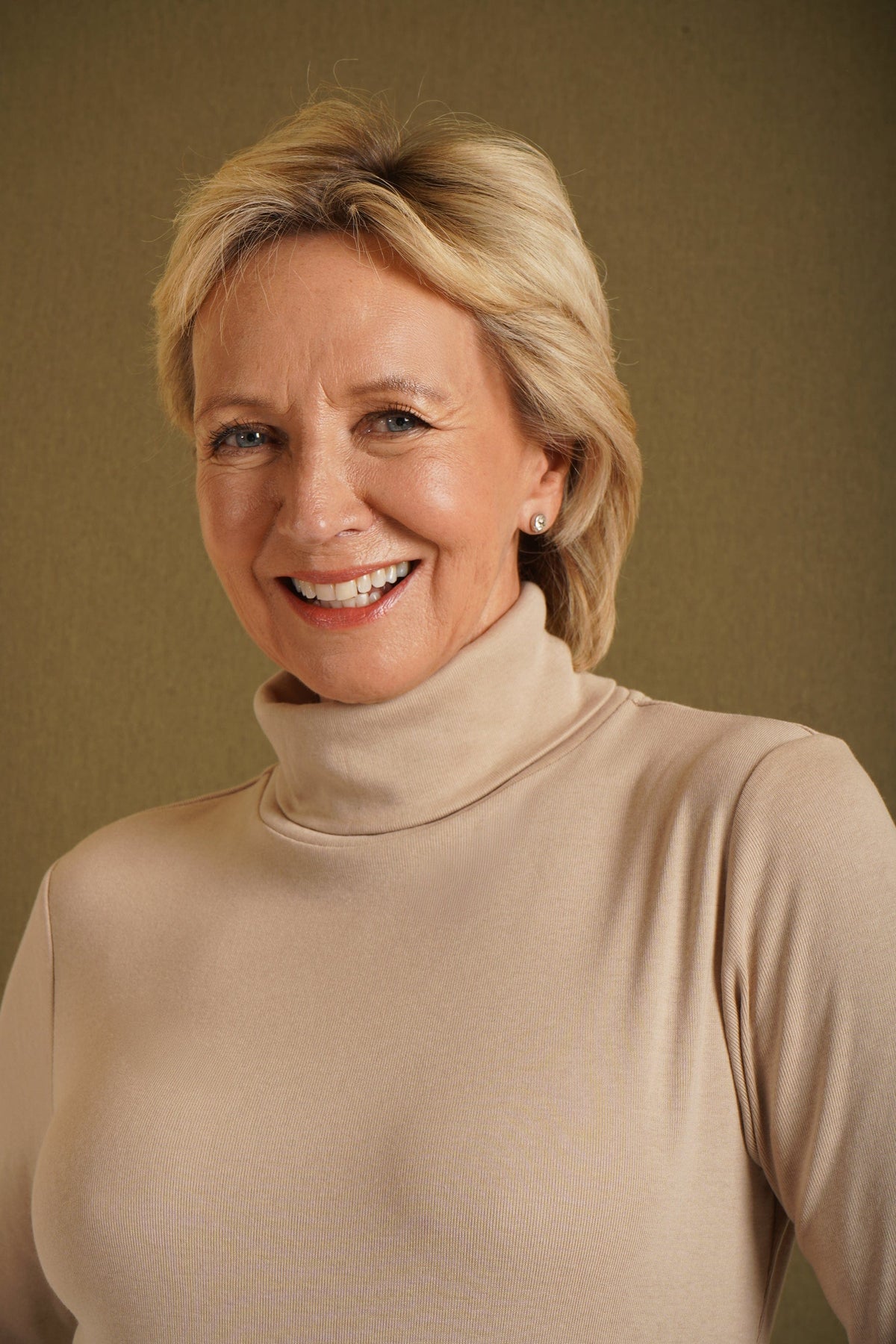 Woman wearing a beige long sleeve turtleneck top with buttoned cuffs and blue jeans against an olive backdrop._6