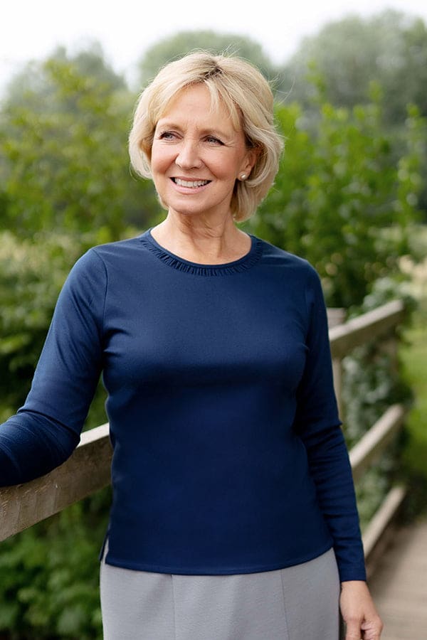 Woman wearing a navy long sleeve top with a ruffle trimmed round neckline and grey trousers, standing on a wooden bridge._0