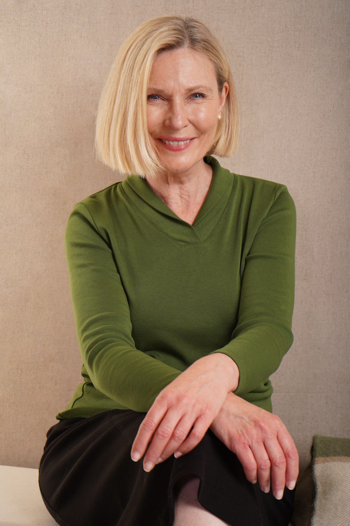Woman with a blonde bob sitting and smiling, wearing an olive green long sleeve v-neck top with ribbed cuffs and a dark brown skirt._0