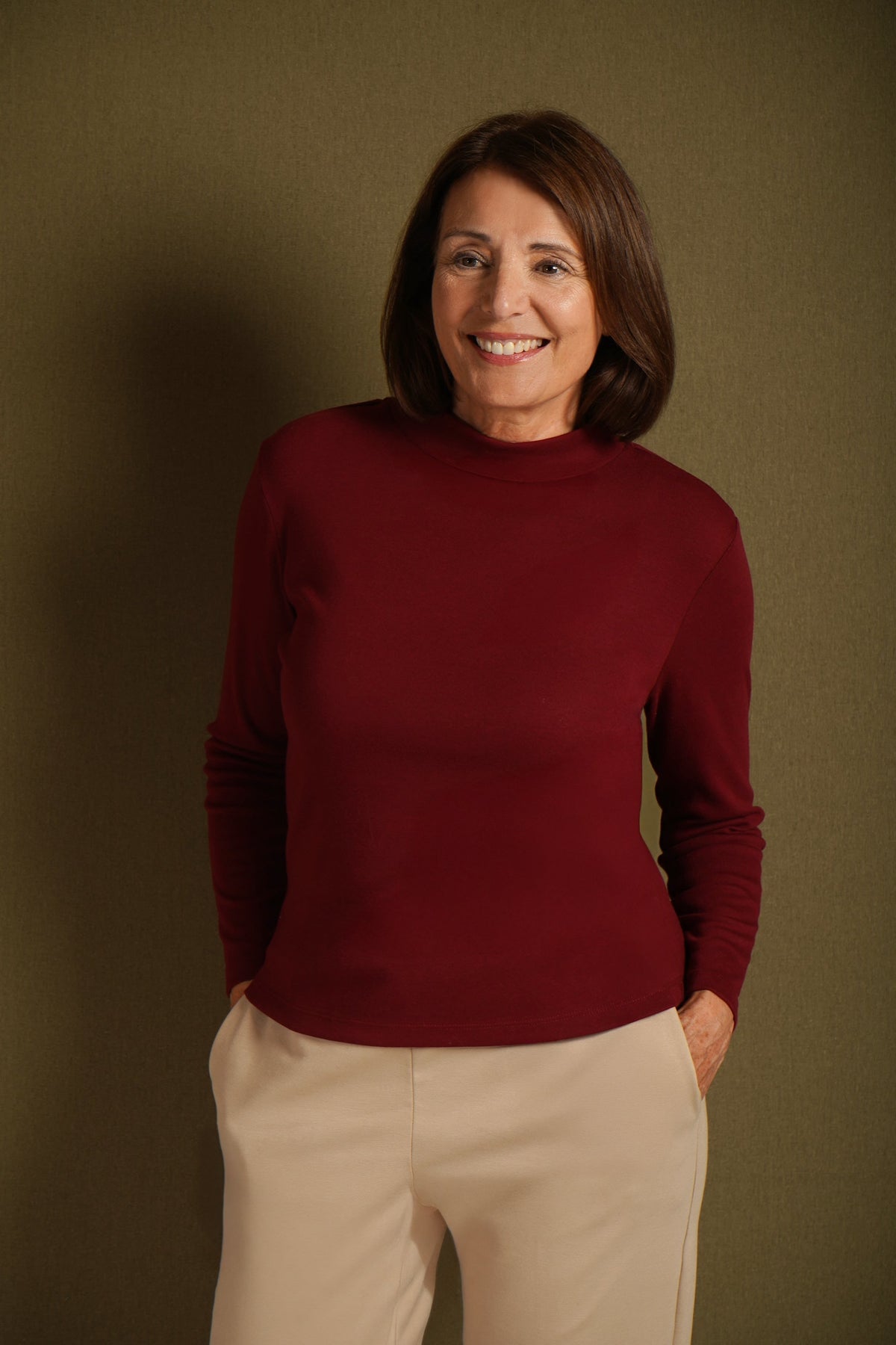Woman wearing a burgundy long sleeve mock neck top with cream trousers against an olive background._0