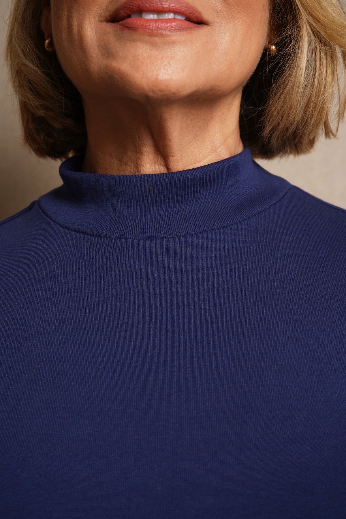 Woman wearing a burgundy long sleeve mock neck top with cream trousers against an olive background._6