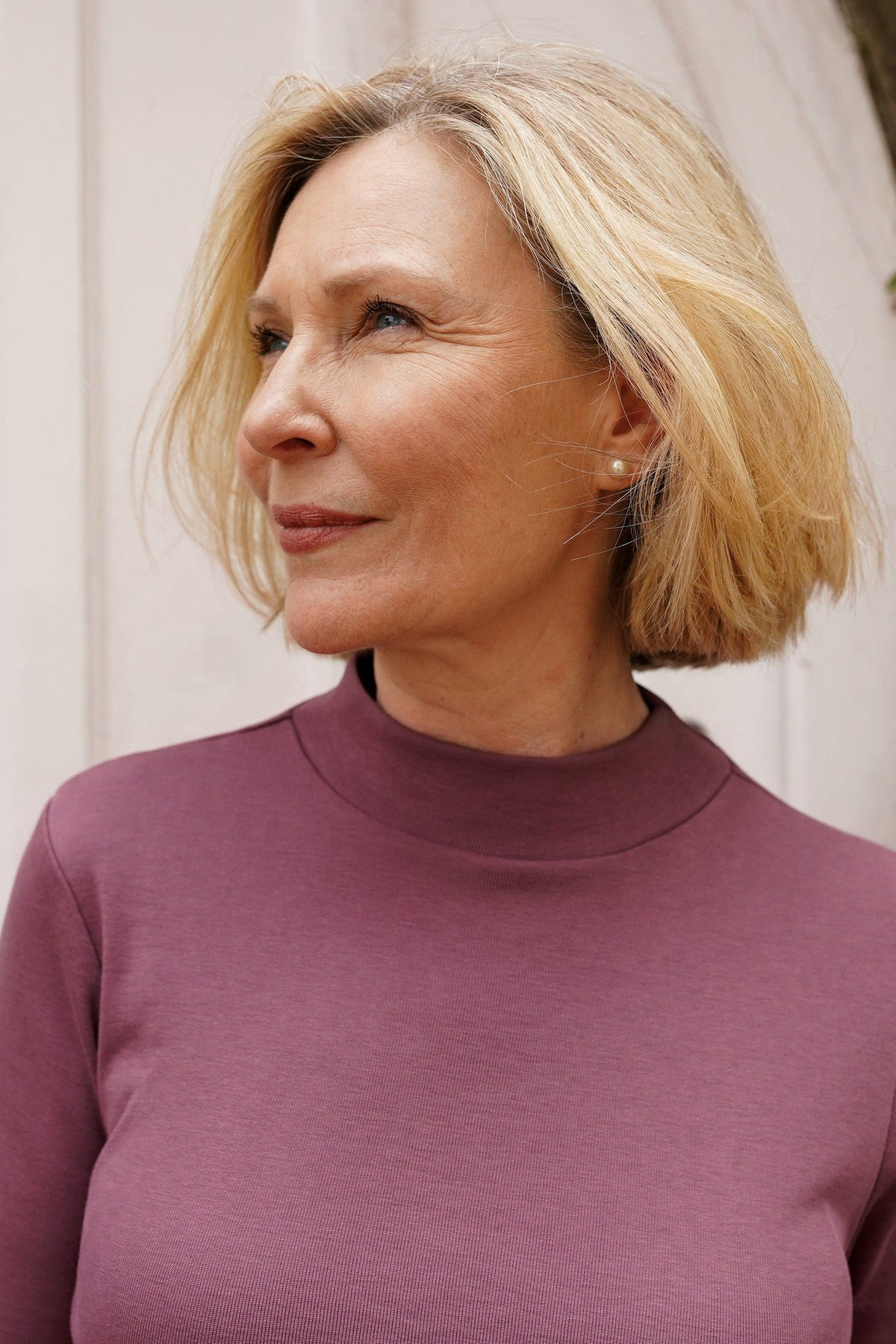 Woman wearing a burgundy long sleeve mock neck top with cream trousers against an olive background._9