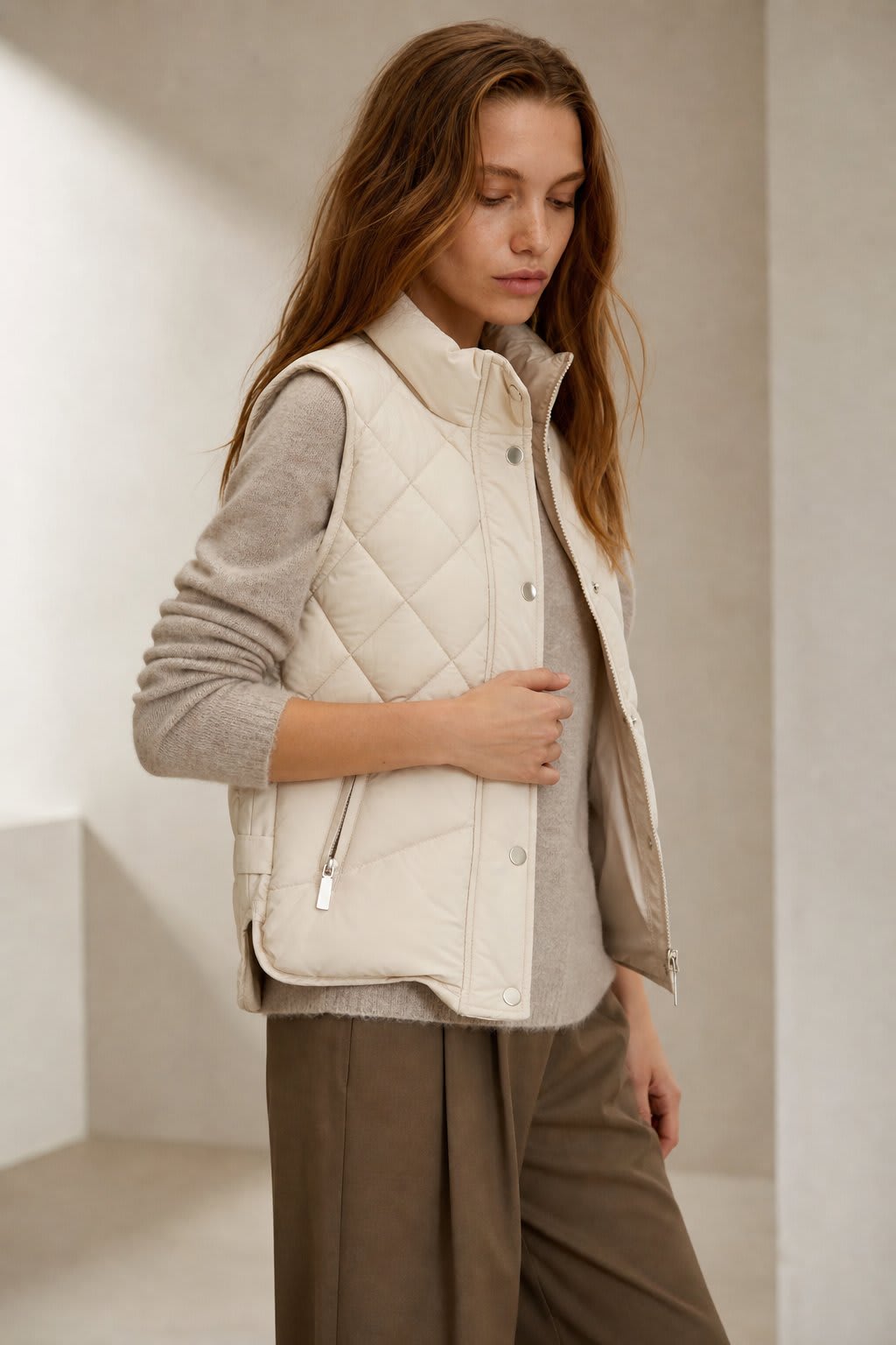 Model wearing a cream quilted vest with stand collar over a neutral sweater and brown trousers._0
