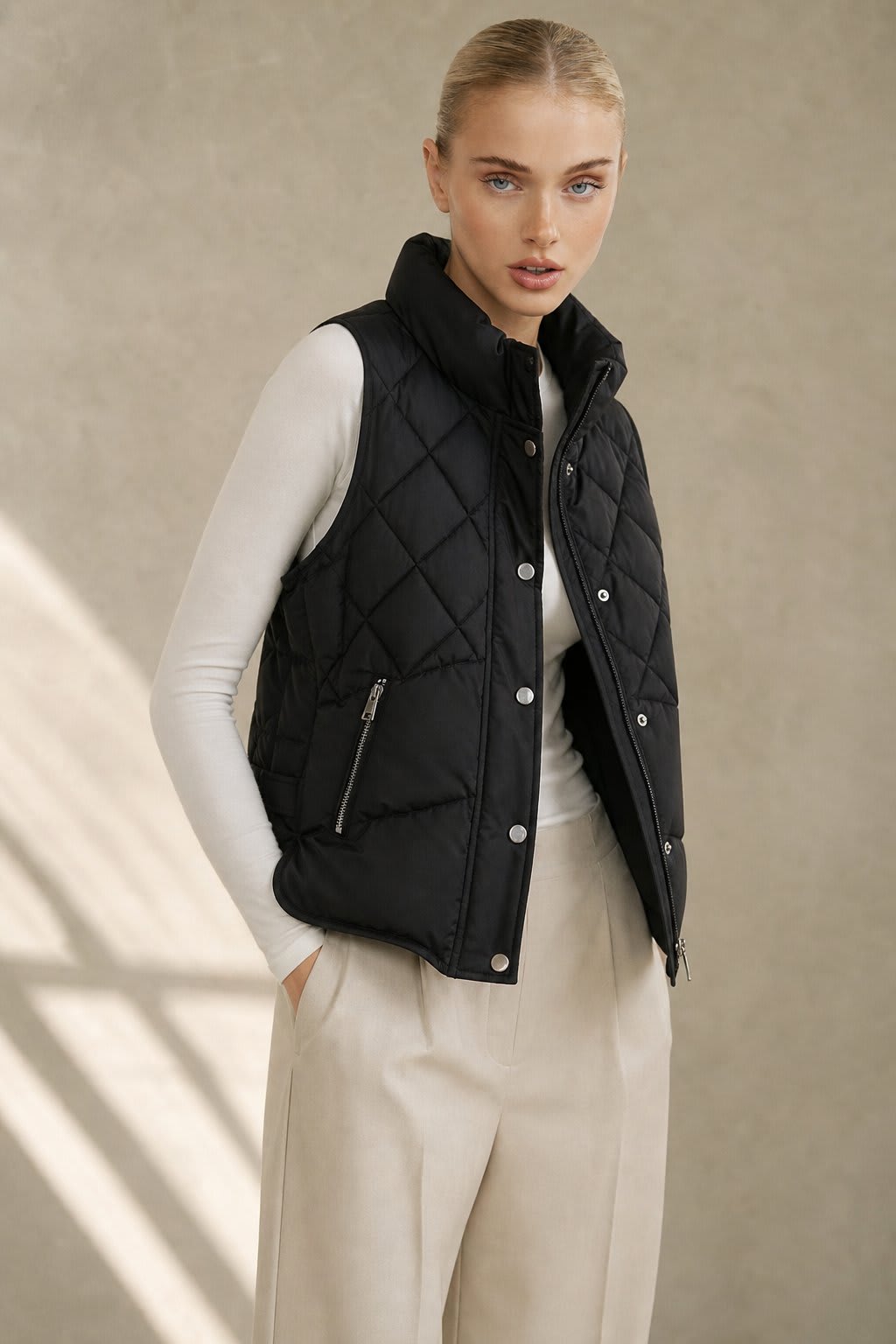 Model wearing a cream quilted vest with stand collar over a neutral sweater and brown trousers._2