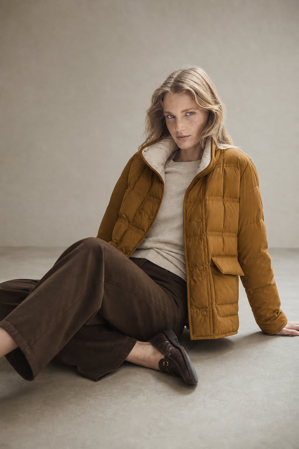 Woman sitting on the floor wearing a mustard quilted jacket with snap button closure over a neutral sweater and brown wide-leg pants._0