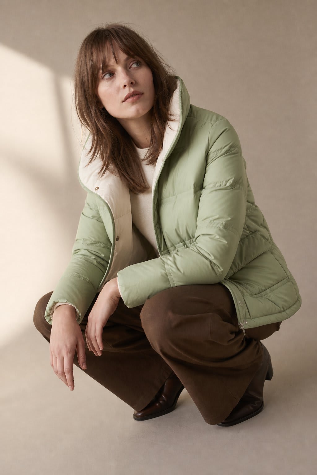 Woman sitting on the floor wearing a mustard quilted jacket with snap button closure over a neutral sweater and brown wide-leg pants._7