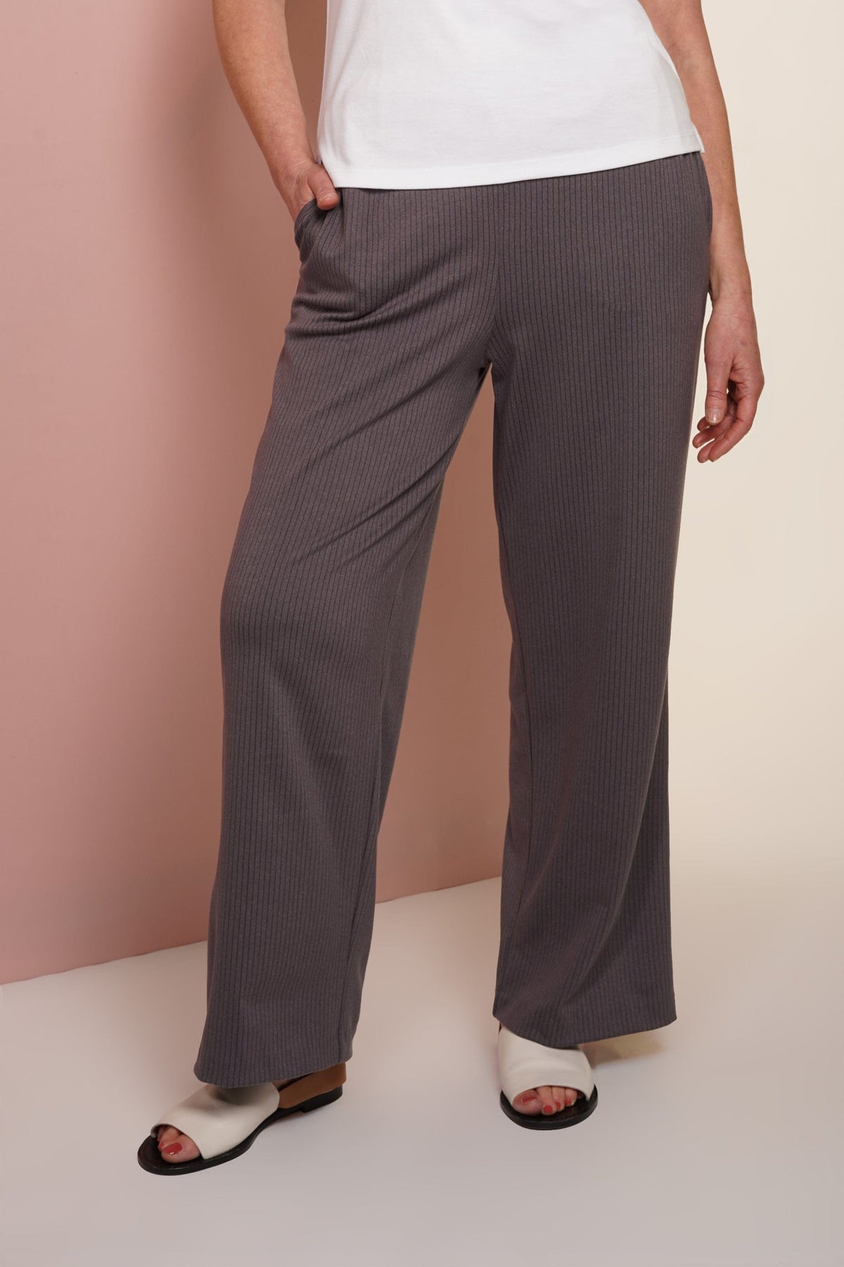 Woman wearing grey ribbed wide-leg trousers with a white t-shirt and white flat sandals against a pink and beige background._0