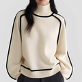 Woman wearing a cream turtleneck sweater with black piping along the raglan sleeves, neckline and ribbed hem, paired with dark trousers._0