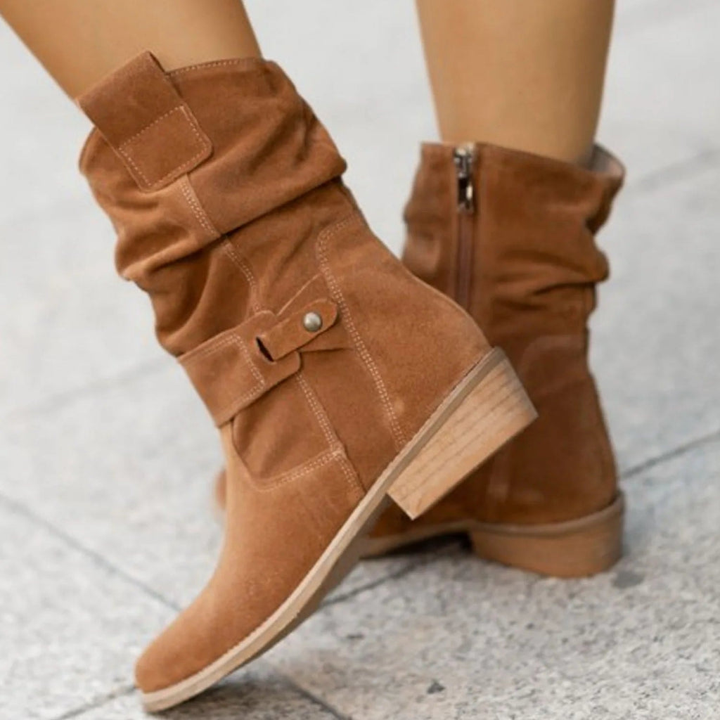 Close side view of tan slouchy boots highlighting the layered shaft, ankle buckle, stitching, and low wood-look heel.