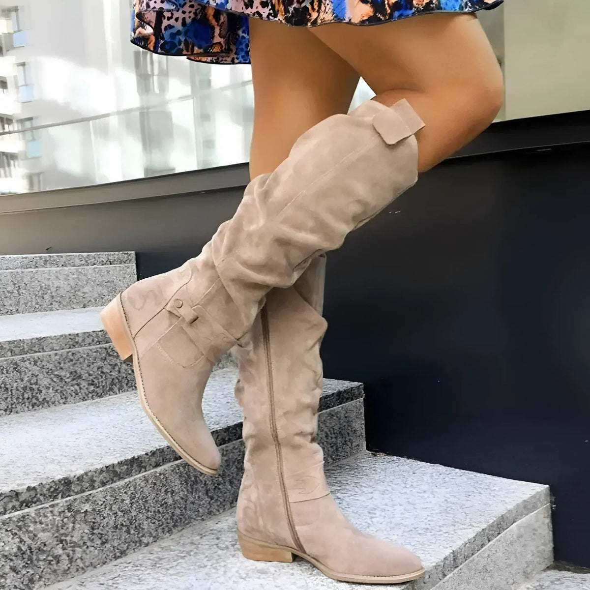 Beige slouchy knee-high boot with a pointed toe and low heel shown on granite steps.