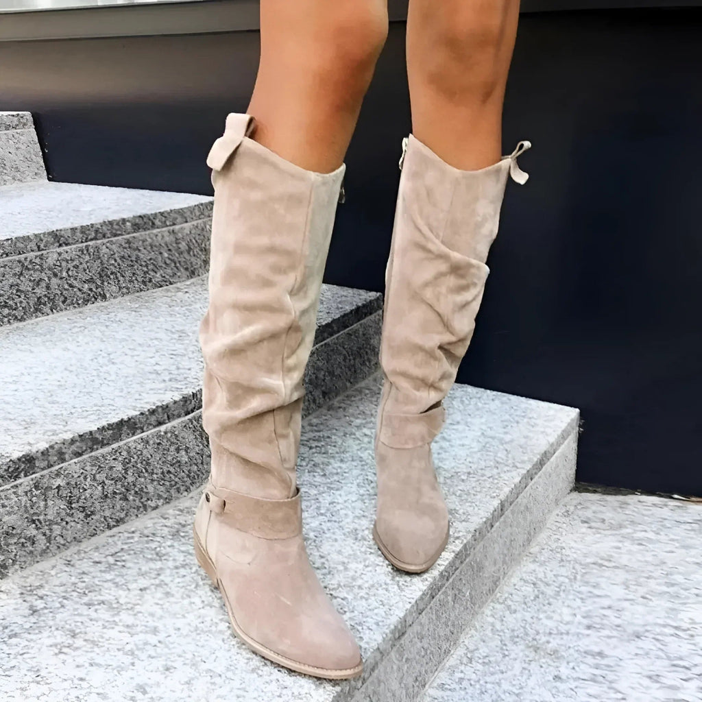 Pair of beige knee-high boots displayed from the front, highlighting their gathered shafts, pointed toes, and low heels.