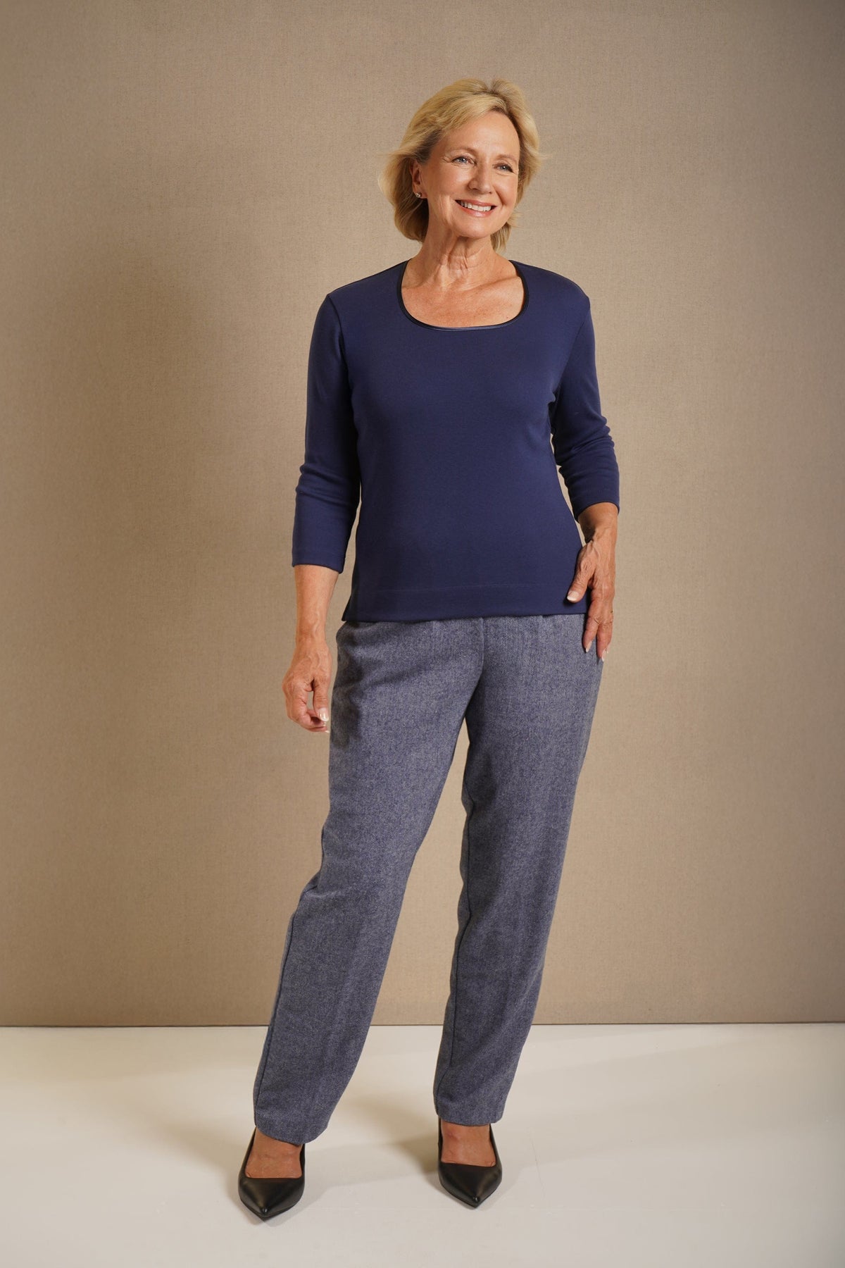 Woman wearing a navy three quarter sleeve scoop neck top with contrast neckline trim, grey trousers and black pointed heels._0