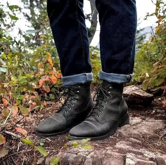 Pair of black lace-up ankle boots with cap-toe detail worn with cuffed jeans outdoors on a wooded trail.
