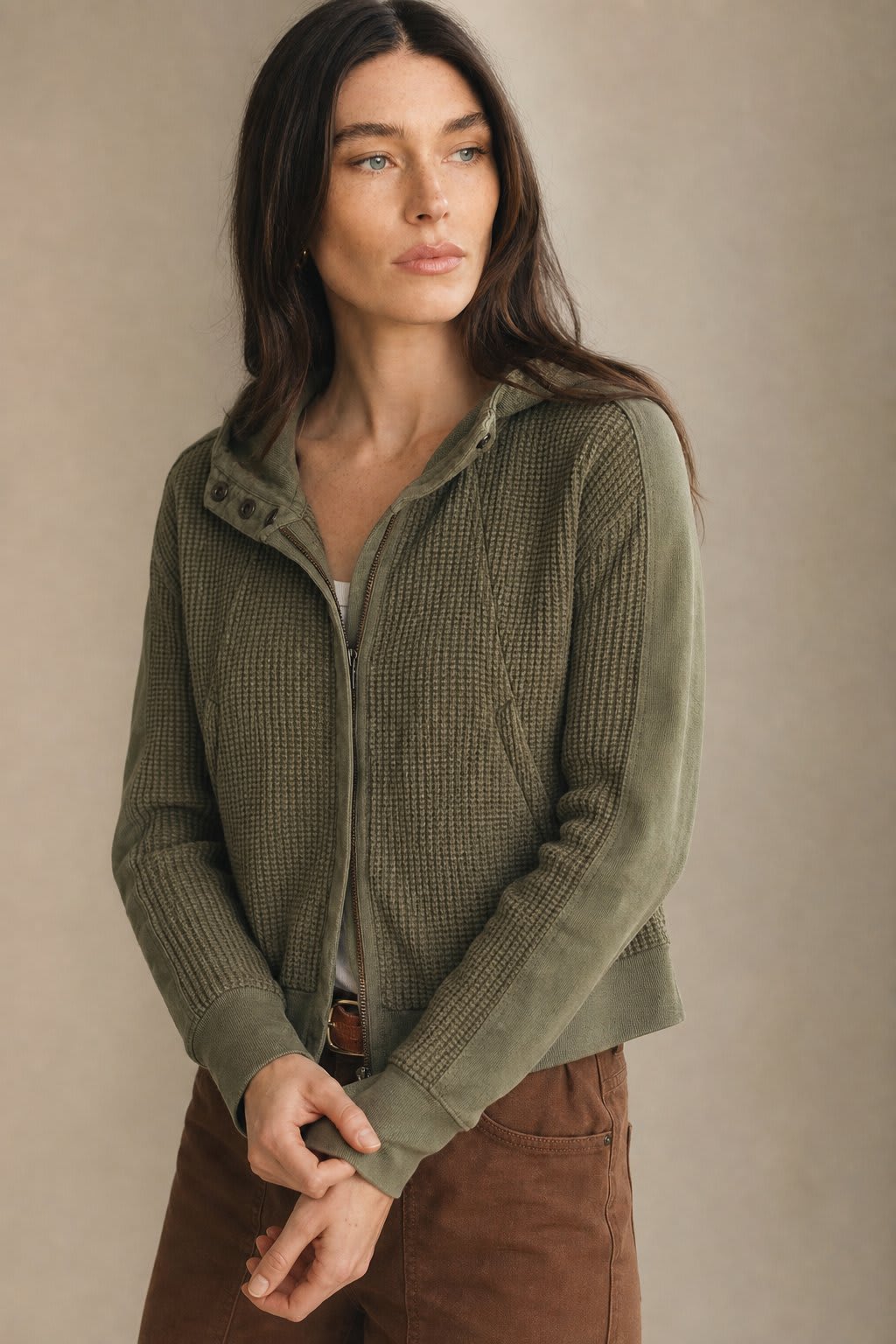 Model wearing an olive green waffle knit zip-up hoodie with cropped length and hood, styled with brown pants._0