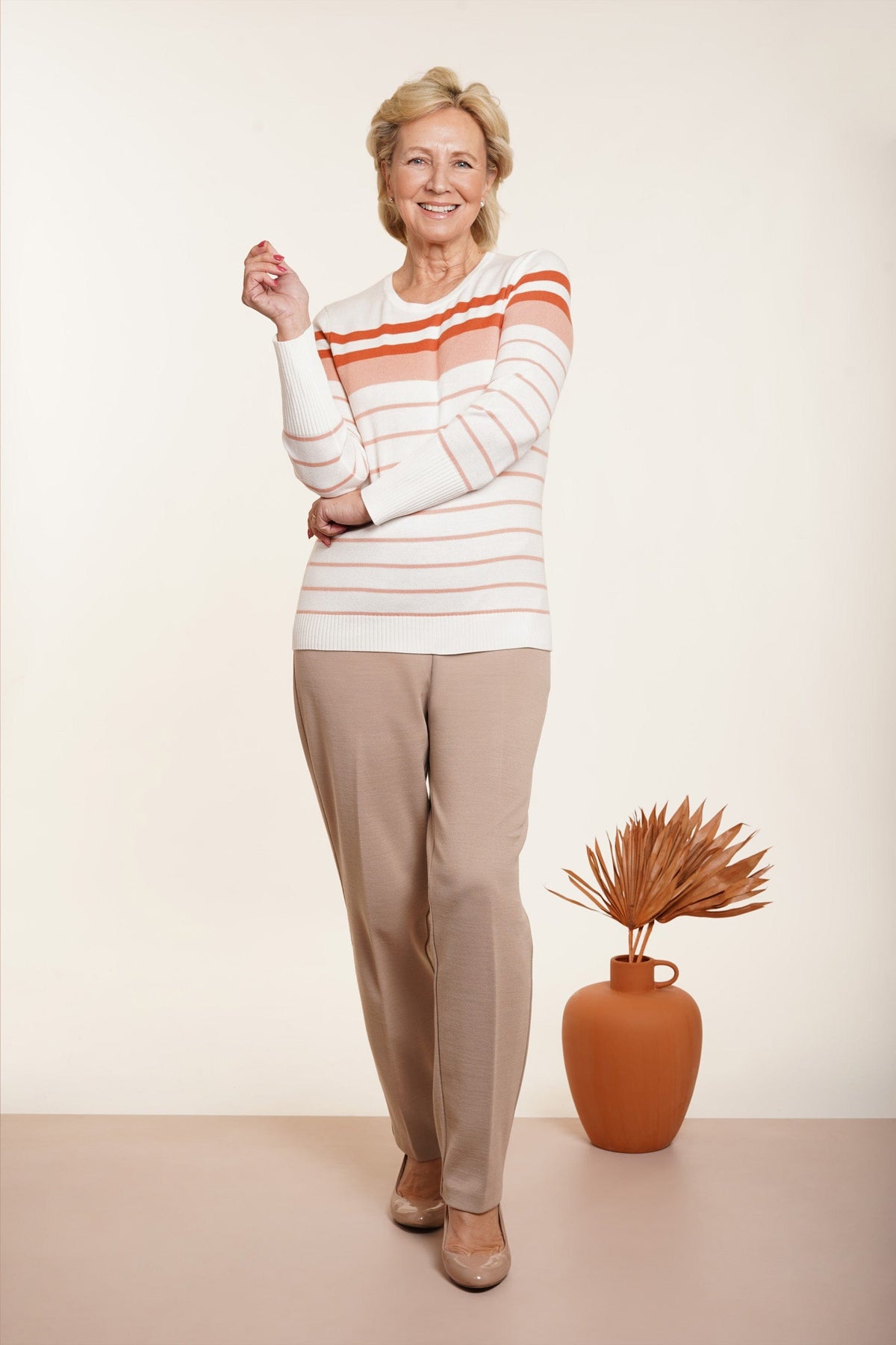 Woman wearing taupe straight leg ankle length trousers with side pockets, a cream striped cardigan and beige braided heeled sandals beside woven baskets._2